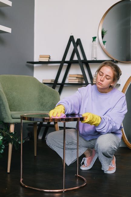 A young woman with light skin and brown hair tied back, dressed in a purple sweatshirt, yellow cleaning gloves, and checkered pants, is kneeling on a dark wooden floor while polishing a round, glass-t