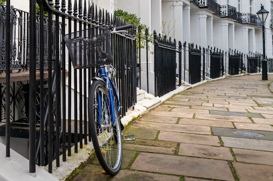An outdoor residential area showing a sidewalk made of large, irregularly shaped stone slabs with visible moss and dirt in the gaps. A black metal fence runs along the edge of a white building, with vertical bars and decorative finials on top. A blue bicycle with a black basket attached to the handlebars is leaning against the fence, positioned near the corner of the building's steps. The building has white columns supporting a balcony, and a black lantern-style street lamp is mounted on the wall. The scene is well-lit with natural daylight, highlighting the clean and well-maintained condition of the surfaces. This setting illustrates aspects of surface cleaning and maintenance, which [COMPANY_NAME] can assist with in the context of domestic and commercial cleaning near West Kensington station.
