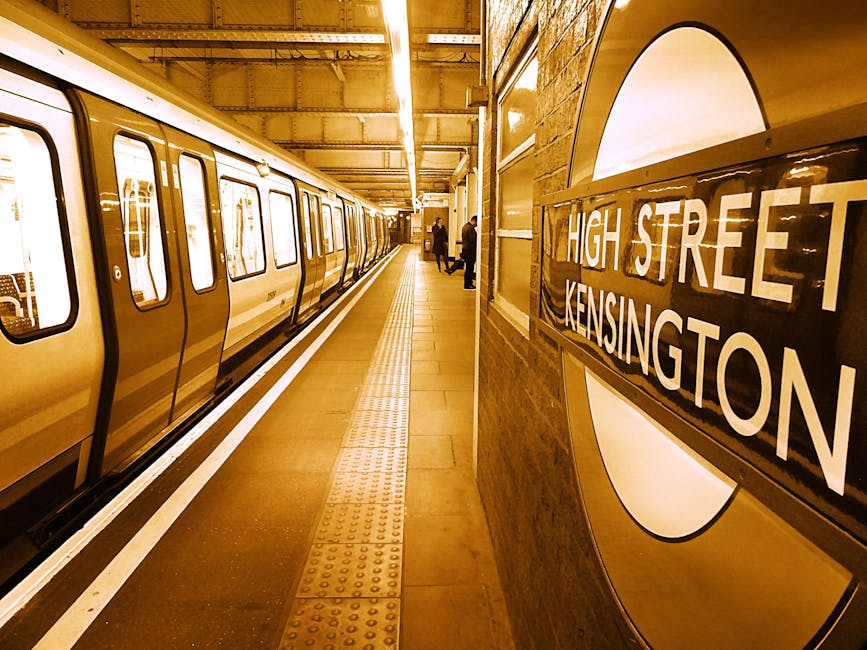 Inside West Kensington underground station showing the platform with a stainless steel train on the left side, illuminated by bright overhead lighting. The platform has a textured yellow tactile strip near the edge for safety, and the floor is clean with smooth tiles. On the right side, there is a brick wall with a large, circular London Underground roundel sign reading 'High Street Kensington'. A few commuters are visible in the distance, either standing or walking along the platform. The station appears well-maintained, with polished surfaces and clear signage, consistent with professional cleaning practices by West Kensington carpet cleaners for maintaining hygiene and surface cleanliness.