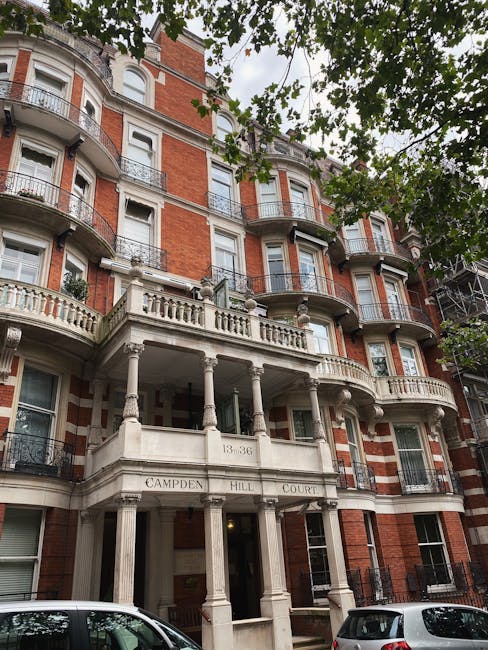 An exterior view of a multi-story residential building at 13-36 Campden Hill Court in West Kensington, constructed with red brick and decorative white stone detailing. The building features curved bay windows with black metal railings on the balconies, and a prominent entrance with classical columns supporting a balcony above. The area is shaded by green leafy trees, and parked cars are visible at the base of the building. The overall appearance is clean and well-maintained, reflecting a tidy and attractive urban residential property, suitable for professional cleaning and maintenance services offered by West Kensington Carpet Cleaners.